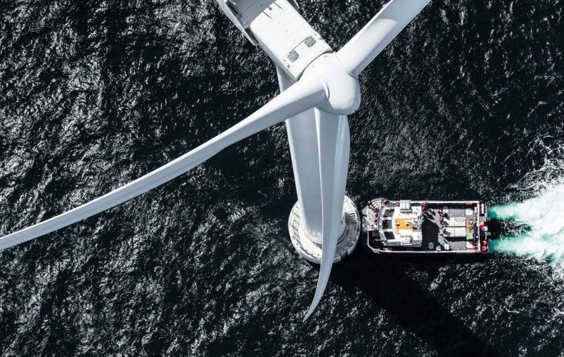 An aerial view of a wind turbine in the ocean, with a support boat positioned near its base.